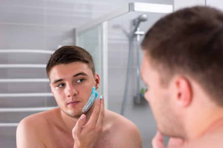 Handsome young man smearing shaving gel on his face standing in front of mirror in the modern tiled bathroom at home